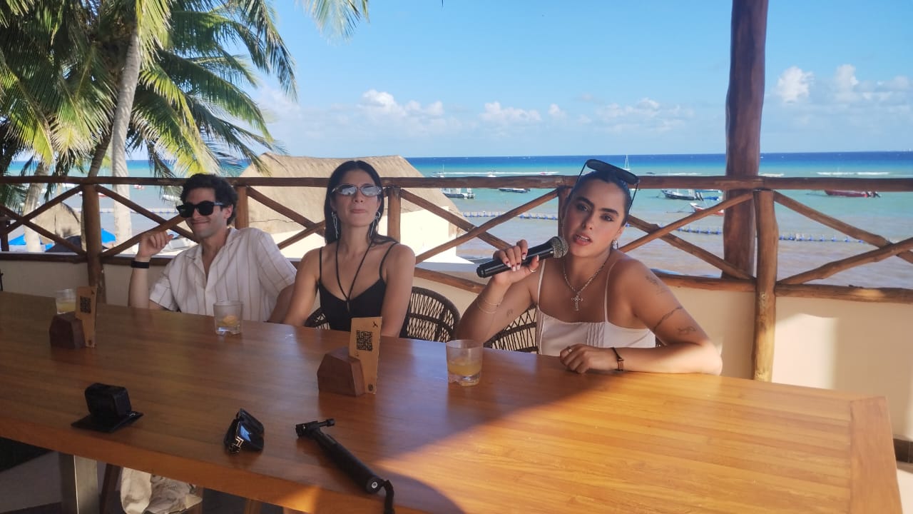 three people sitting at a wooden table with drinks in a beachside setting 27102025