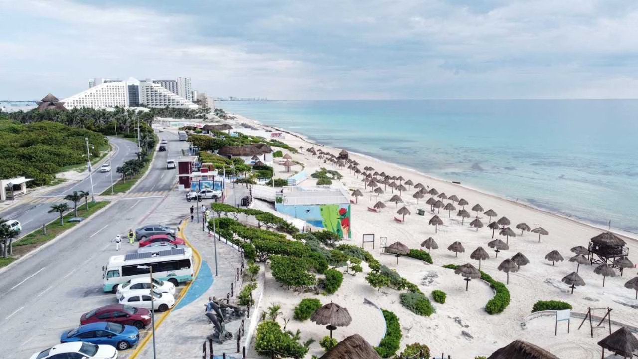 aerial view of a beach with straw huts and a road lined with cars and greenery-21102025
