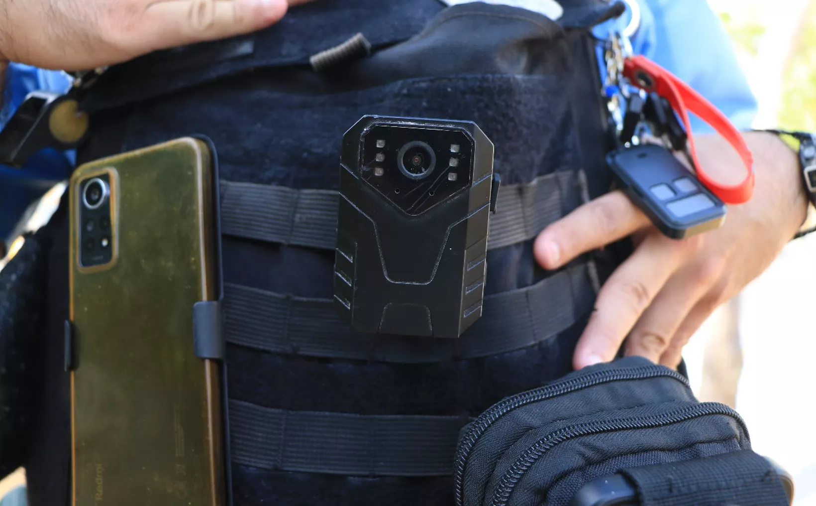 Close-up of a tactical vest featuring a body camera attached, alongside a mobile phone and keychain.$# CAPTION