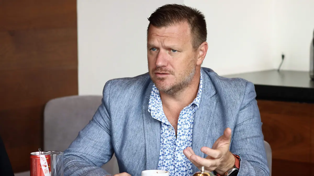 A man in a blue blazer and patterned shirt engaged in a conversation at a table with drinks.$# CAPTION