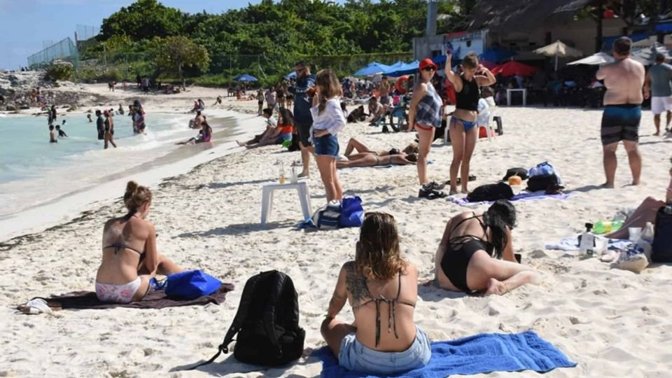 A busy beach with people sunbathing and enjoying the water on a sunny day. An assortment of beach towels, umbrellas, and relaxed vacationers can be seen.