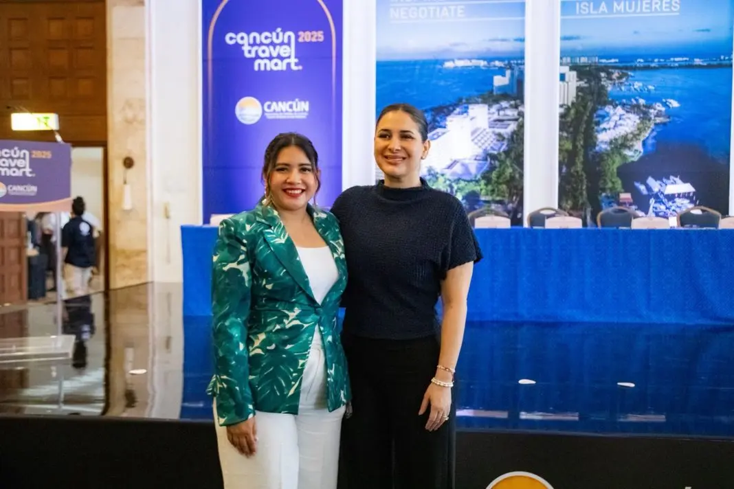 Two women posing for a photo at a conference event with banners behind them in Cancun, Mexico. The woman on the left is wearing a green patterned blazer and white trousers, while the woman on the right is in a black top and pants.$# CAPTION