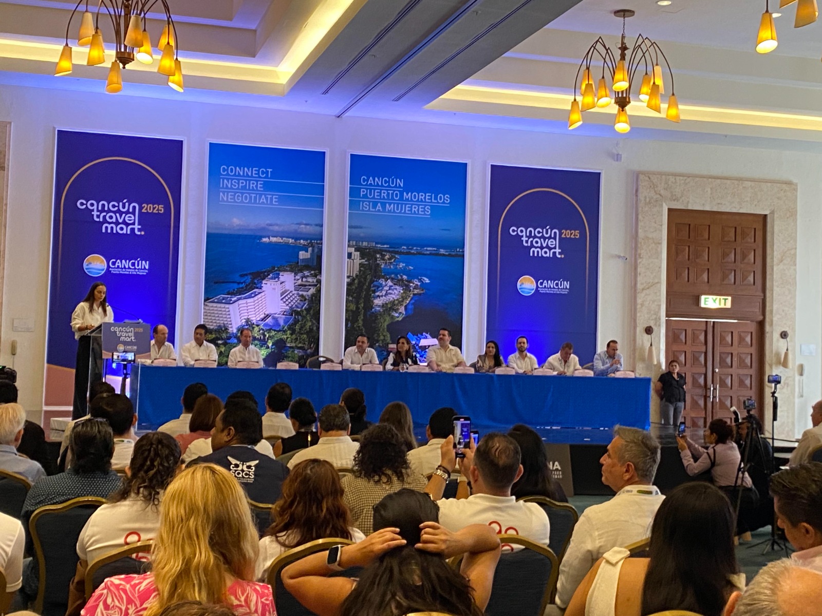 A speaker addresses an audience during the Cancun Travel Mart 2025 event, with a panel of experts in the background.$# CAPTION