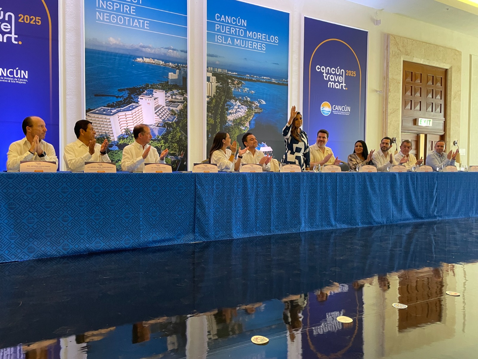 A group of individuals sitting at a table during a press conference for Cancun Travel Mart 2025, with banners in the background portraying scenic views of Cancun.$# CAPTION