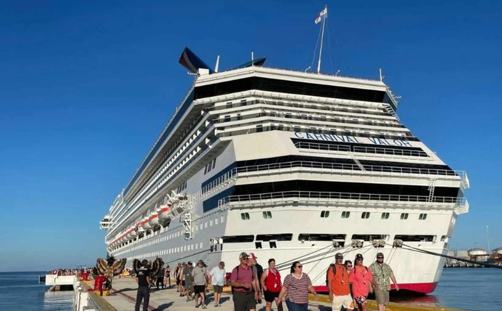 A large cruise ship, the Carnival Valor, docked at a port with passengers walking on the dock in the foreground under a clear blue sky.$# CAPTION