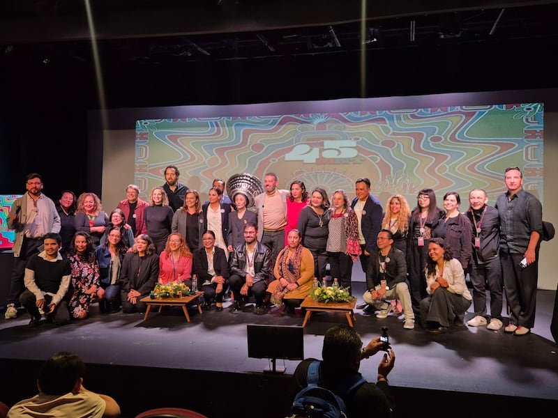 A large group of people, including men and women, gathered on stage for a photo opportunity during a celebratory event, with a colorful backdrop and floral arrangements in front of them.$#$ CAPTION