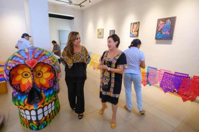 Two women converse in an art gallery decorated with colorful papel picado and a vibrant skull sculpture, while other visitors admire the artwork on the walls.$