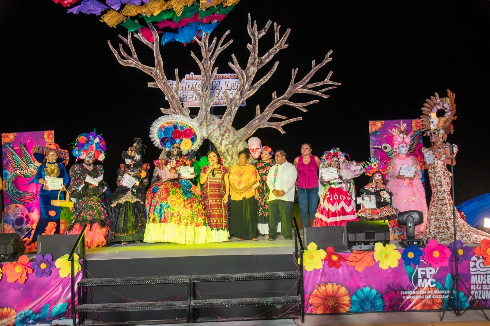 A group of participants in colorful traditional costumes on stage during a Day of the Dead celebration, with vibrant decorations and a large tree in the background.$# CAPTION