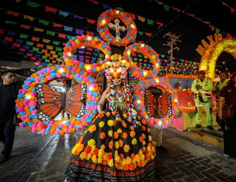 A performer dressed in a vibrant costume adorned with floral decorations and skull motifs, standing in a colorful celebration backdrop for Día de los Muertos, surrounded by festive decorations and people.$# CAPTION