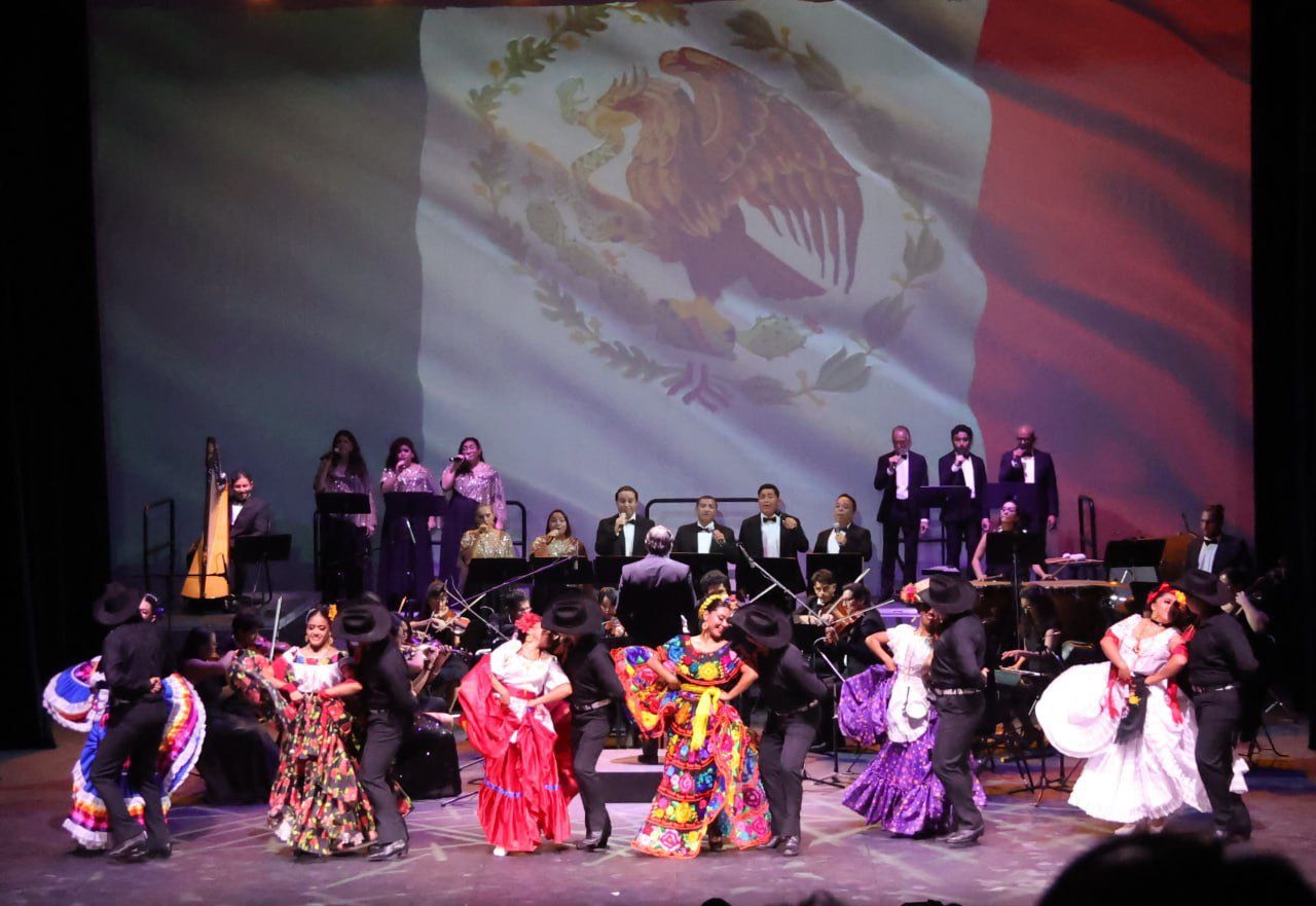 A vibrant dance performance featuring traditional Mexican dancers in colorful costumes, with musicians and a large Mexican flag backdrop.$# CAPTION
