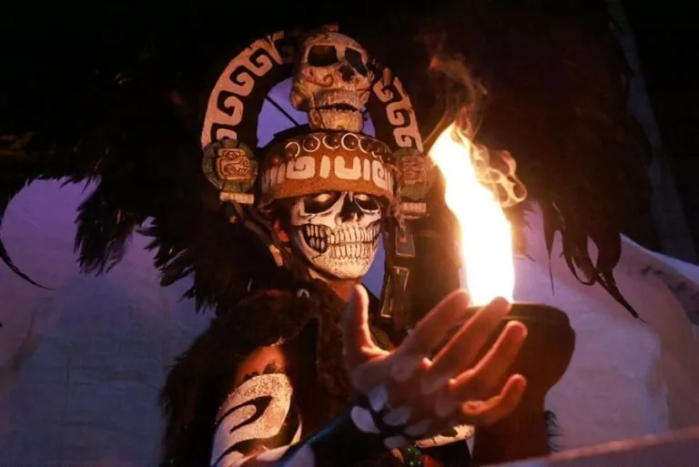 A person in traditional attire with face paint resembling a skull holds a bowl emitting flames.$# CAPTION