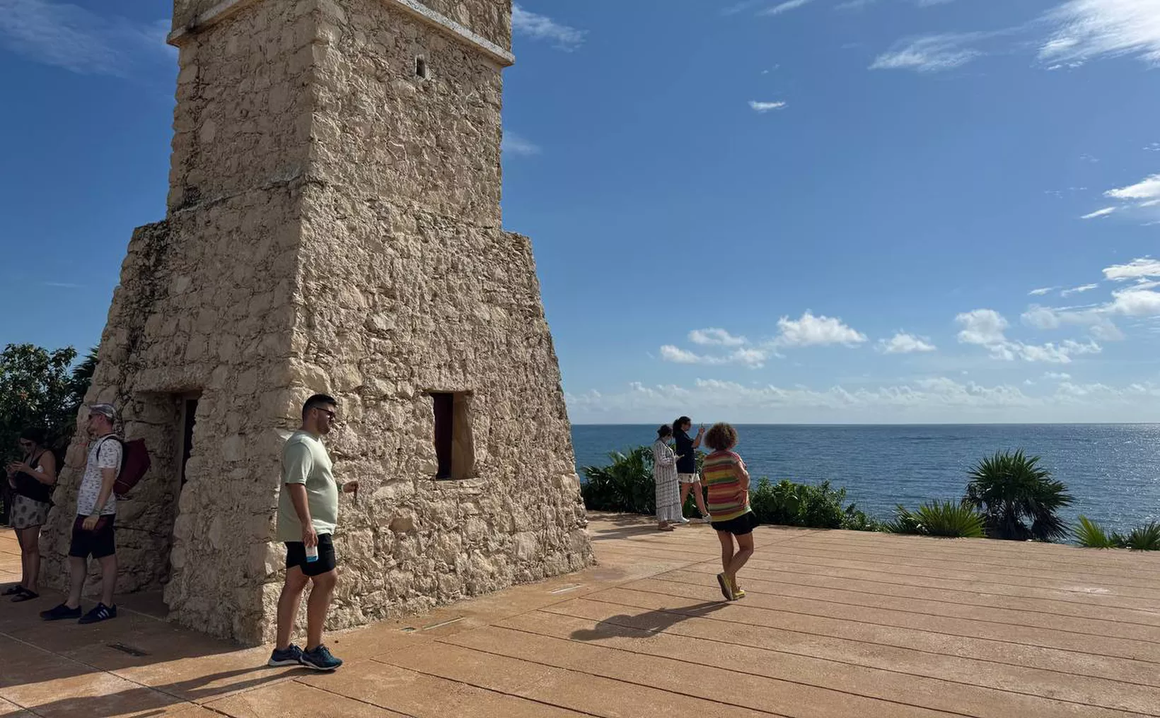 A group of people exploring ancient stone ruins by the ocean on a sunny day with blue skies and scattered clouds.$#$ CAPTION