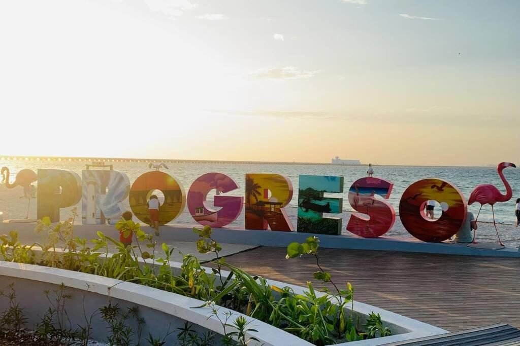 A vibrant sign spelling "PROGRESO" with palm trees and flamingos, set against a sunset over the ocean.$
