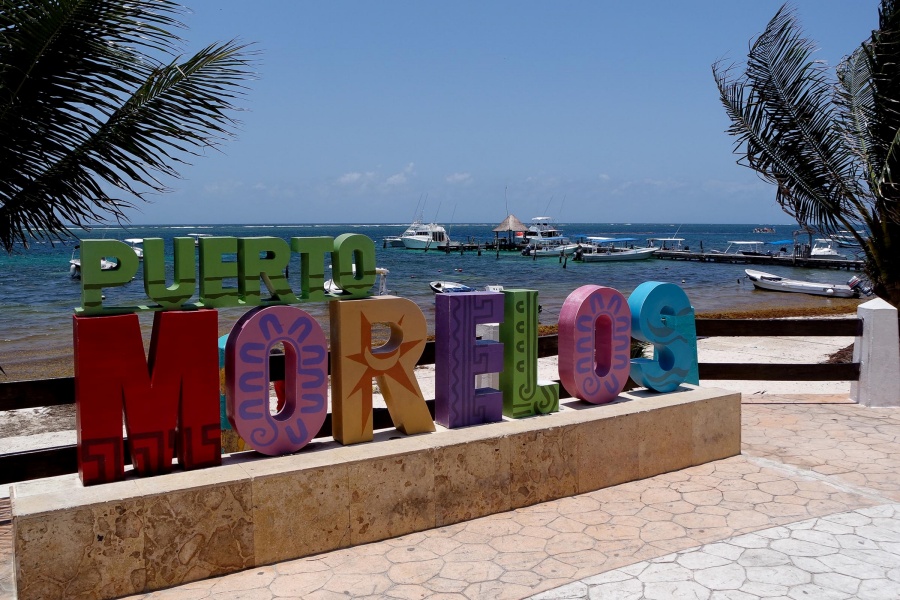 A vibrant sign reading "Puerto Morelos" in various colors, set against a tropical beachfront with palm trees and boats in the background.$# CAPTION