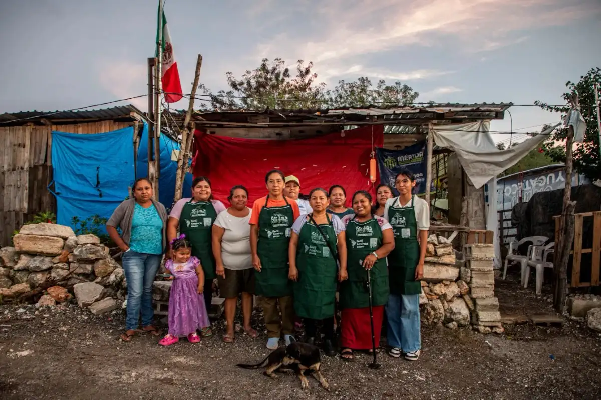 A group of women wearing green aprons stand together in front of a makeshift building, with a small girl in a purple dress and a dog at their feet. Mexican flags and colorful tarps are visible. $#$ CAPTION