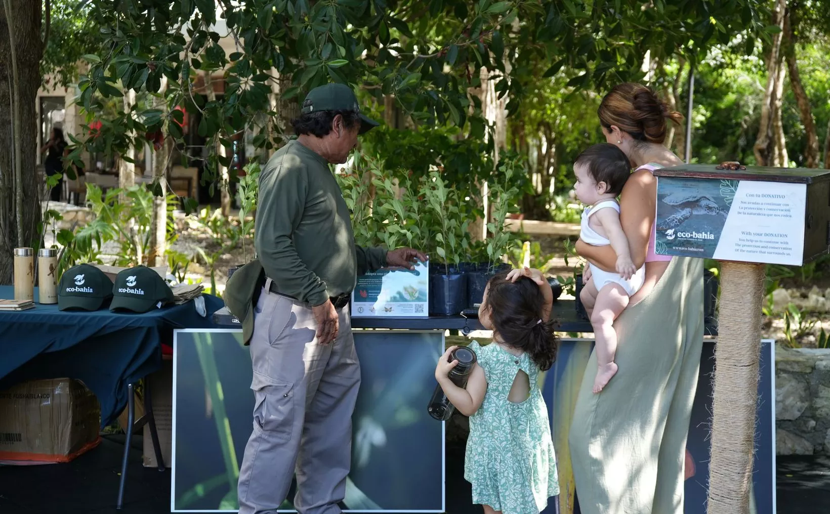 A group of people, including an adult man and two children, interacting with a display of plants and educational materials at Eco-Bahia, a conservation initiative.