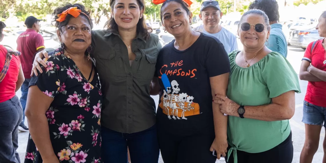 A group of four women posing together at a community event, smiling and showing a vibrant atmosphere. One woman wears a floral dress, another a Simpsons t-shirt, and two have colorful bows in their hair. Background features other attendees and parked cars.