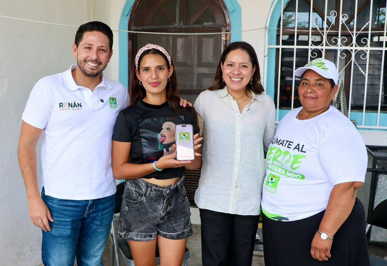 Four individuals stand together, smiling, with one person holding a smartphone displaying a green logo.$# CAPTION