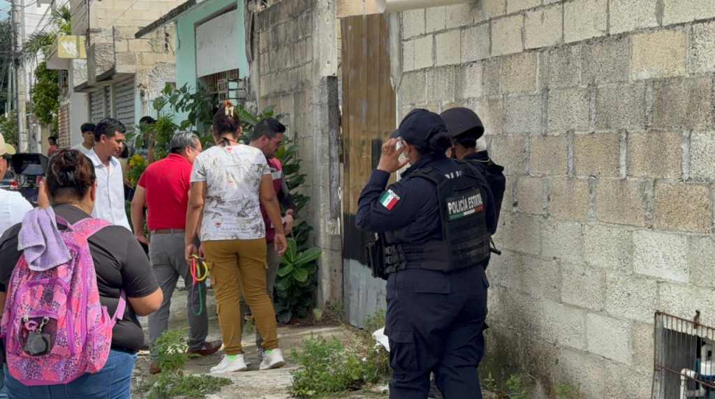 A group of people gathered near a police presence outside a building in a residential area. Two police officers in tactical gear are watching the scene while community members interact nearby.
