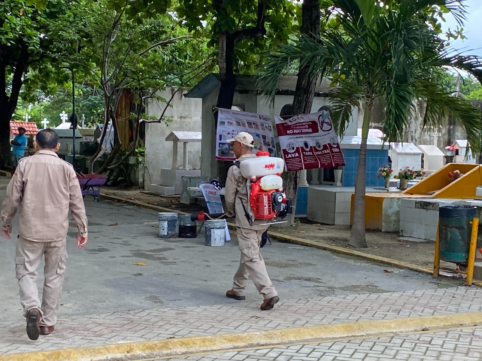 Two individuals walking in a cemetery, one carrying a backpack sprayer for pest control or sanitization, with signs in the background.$# CAPTION