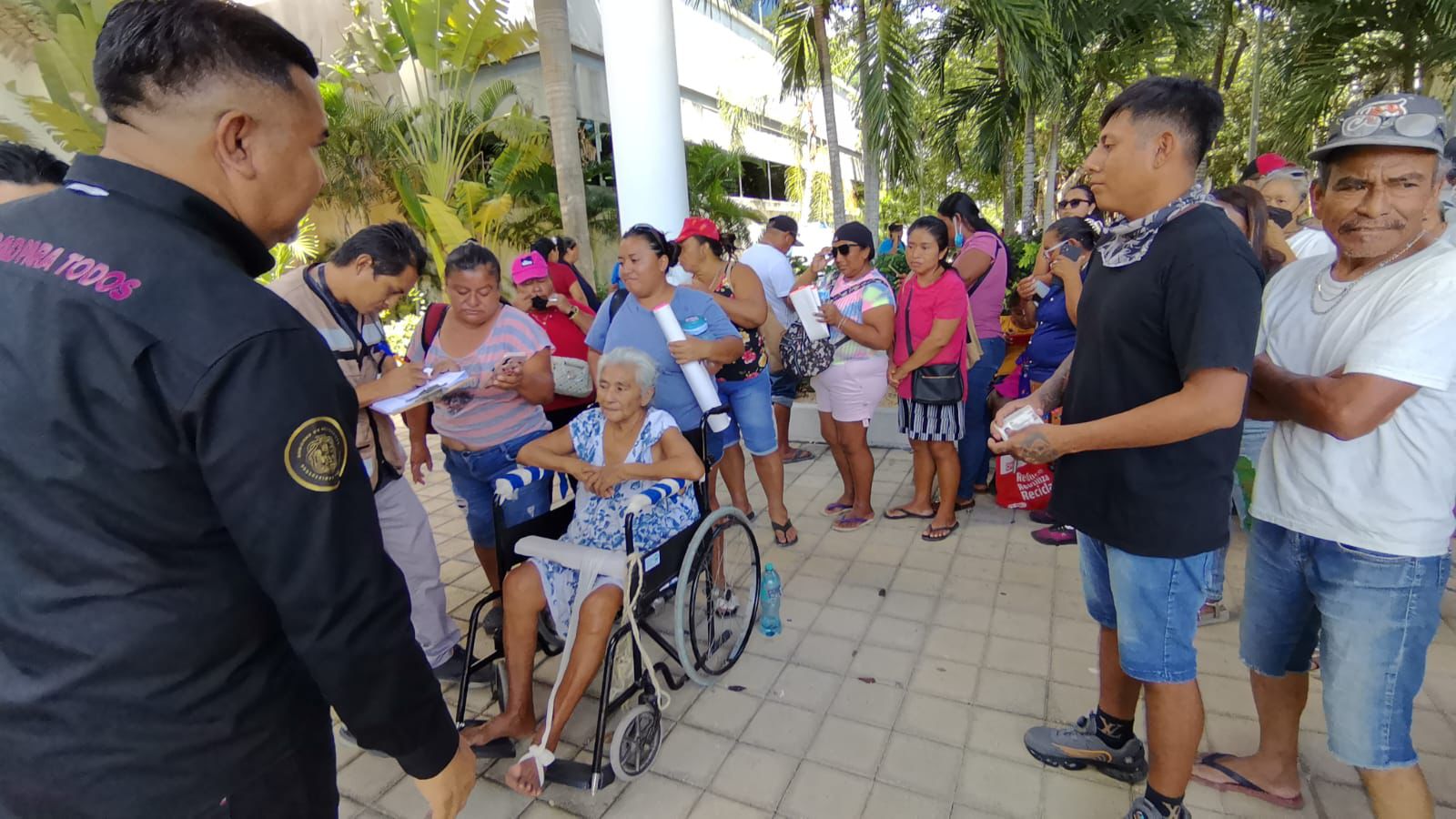 A group of people, including an elderly woman in a wheelchair, standing in line at a community event. Various individuals are interacting, with some holding papers and others with bags. The setting is outdoors with greenery in the background. $#** **CAPTION