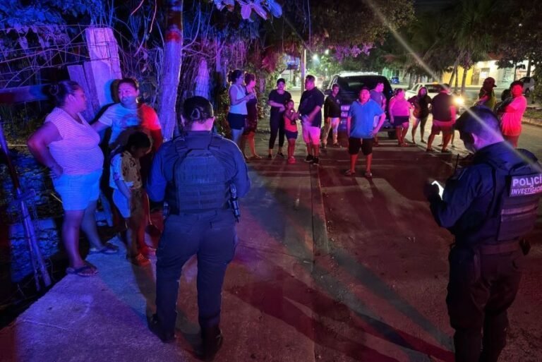 A group of people gathered on a street at night, with police officers overseeing the scene. The atmosphere is tense as various individuals engage in conversation while emergency lights illuminate the surroundings.