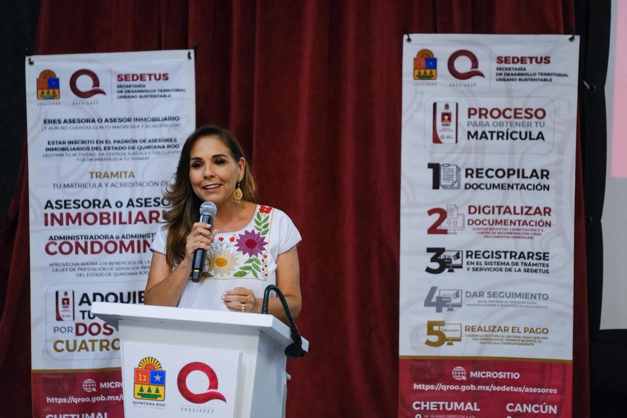 A woman speaking at a podium during a real estate workshop, with informational banners in the background about registration and documentation processes.