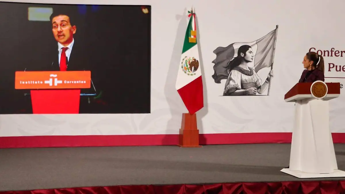 A speaker presenting at a conference in Mexico, with a large screen behind displaying their image, alongside the Mexican flag and a prominent artwork.$#$ CAPTION