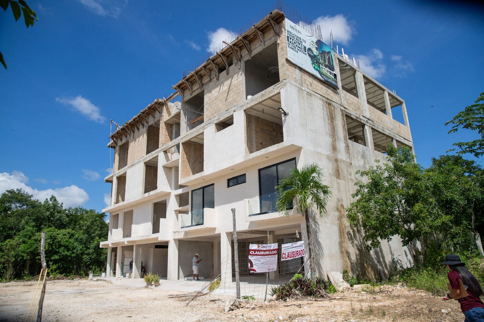 An unfinished multi-story building with large windows, surrounded by greenery and signs indicating closure.