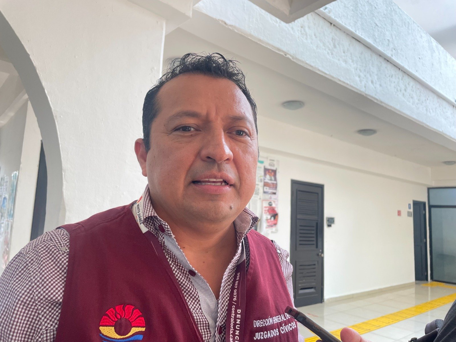 A man speaking in a government office setting, wearing a reddish-brown vest with an emblem, with a white wall and tiled floor in the background