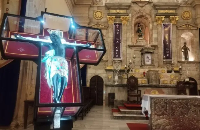 A crucifix displayed inside a church, featuring a statue of Jesus on the cross with an ornate altar in the background.$# CAPTION