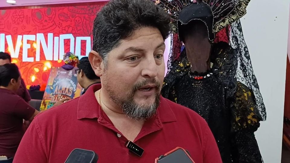 a man in a red shirt speaks during a cultural event with decorations in the background and a traditional costume nearby