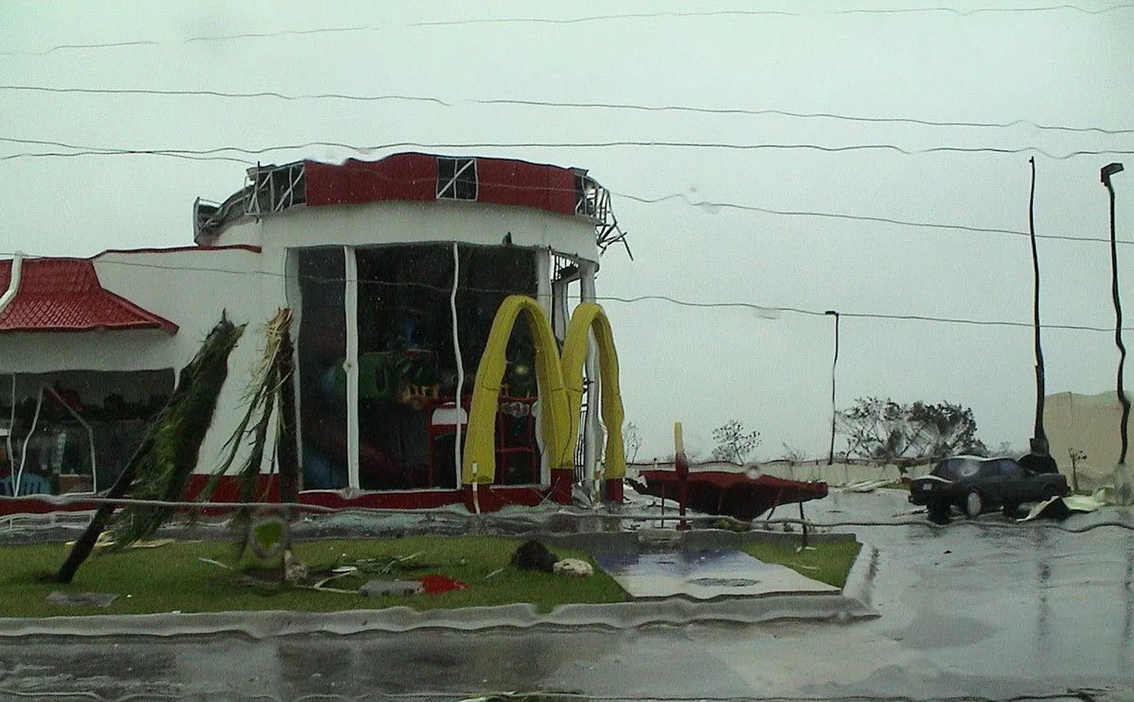 A heavily damaged McDonald's restaurant after a natural disaster, with debris and bent trees visible in the foreground and rain-soaked surroundings.