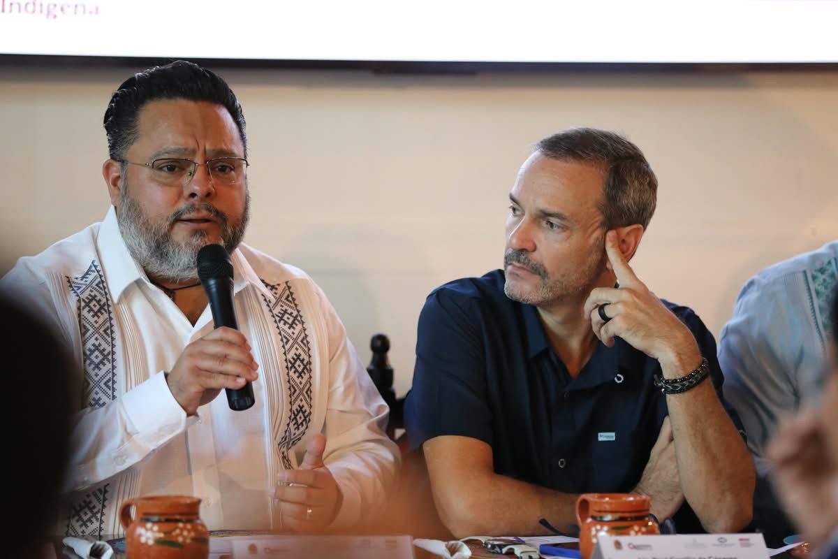 A man with glasses speaking into a microphone at a conference table, beside another man listening attentively. Decorative pottery is in the foreground.$# CAPTION