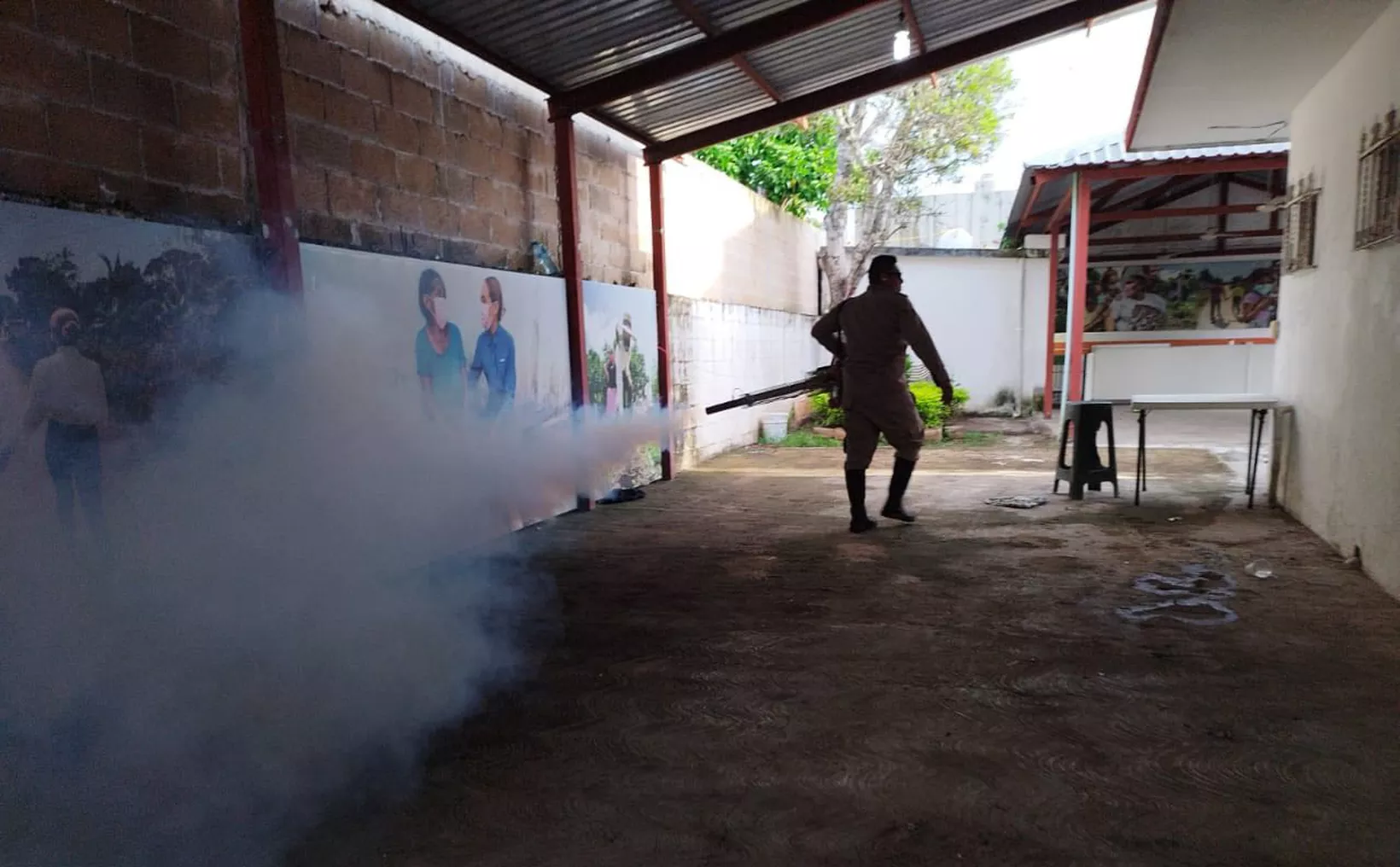 A person in protective gear spraying disinfectant in a well-lit area with murals on the walls, surrounded by fog from the spray.$# CAPTION