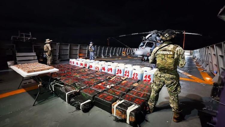 Military personnel inspect a large cache of seized narcotics on a naval vessel at night, with a helicopter in the background.$# CAPTION