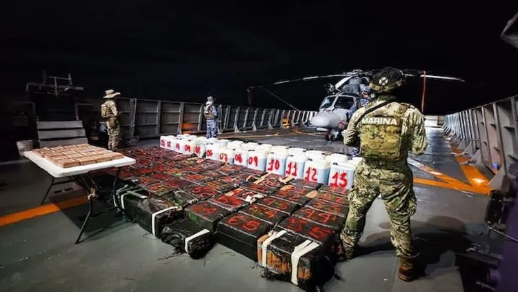 Military personnel inspect a large cache of seized narcotics on a naval vessel at night, with a helicopter in the background.$# CAPTION