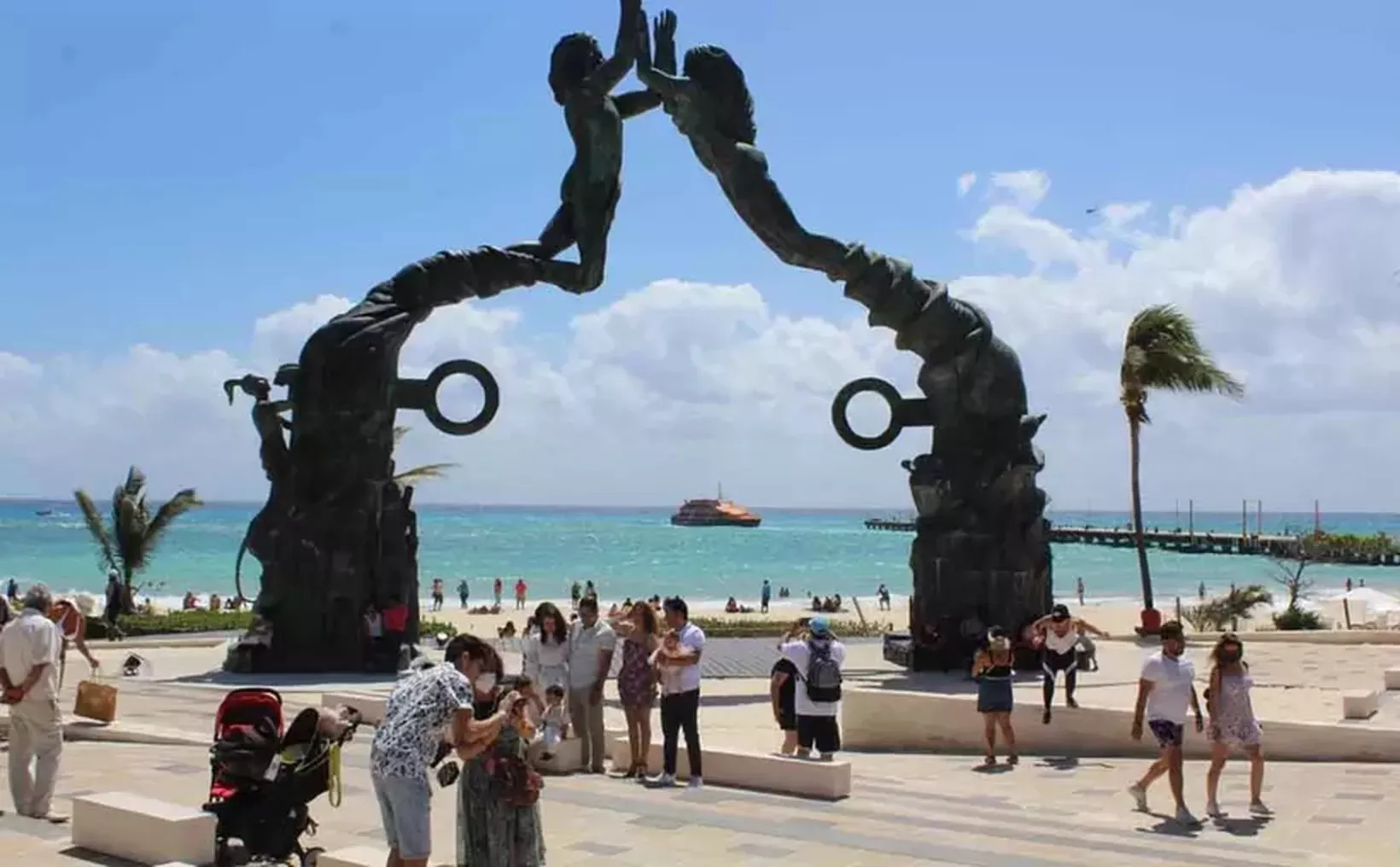 A large sculpture of two figures reaching out to each other with the ocean and beach in the background, surrounded by people enjoying the area.