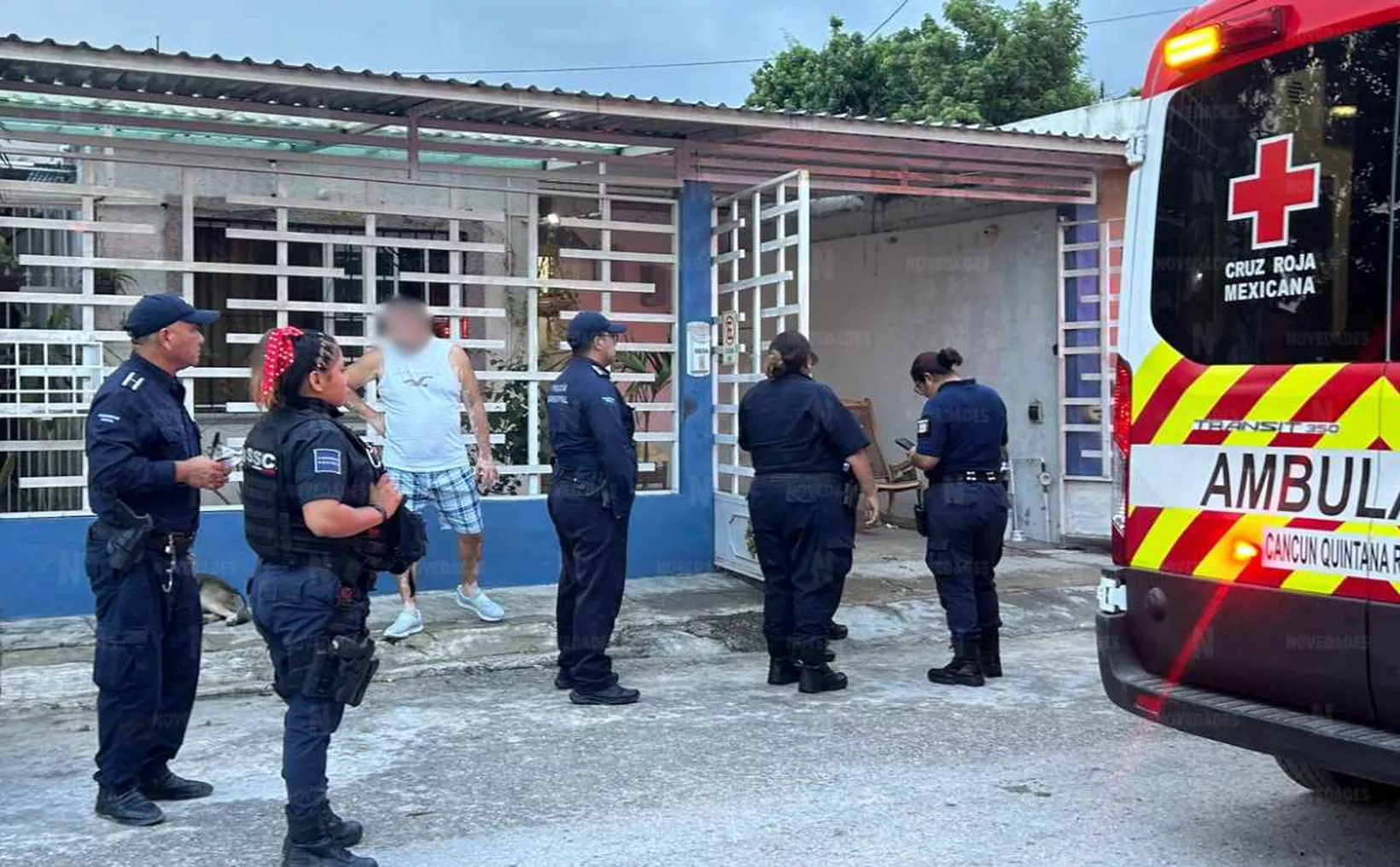 A group of police officers and a paramedic near a residential area in Cancun, Mexico, responding to an incident. A man stands outside the house wearing plaid shorts and a tank top. An ambulance is parked nearby.