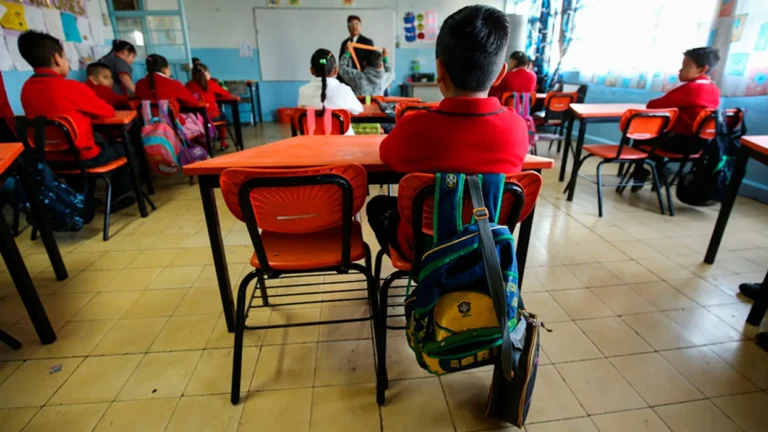 A classroom filled with students sitting at desks, attentively looking at the teacher in front of them. Most students are wearing red uniforms, and one student has a backpack beside them. The classroom has colorful decorations and a whiteboard at the front.