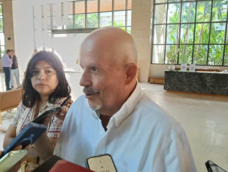 A man in a white shirt speaks while surrounded by microphones, with a woman listening attentively in the background.$# CAPTION