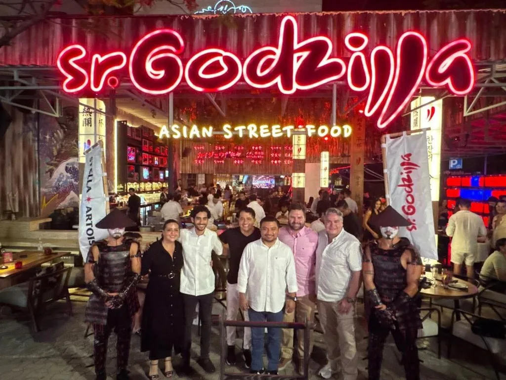 Group of people posing in front of the Sr. Godzilla restaurant with neon signage and entertainers dressed in traditional attire