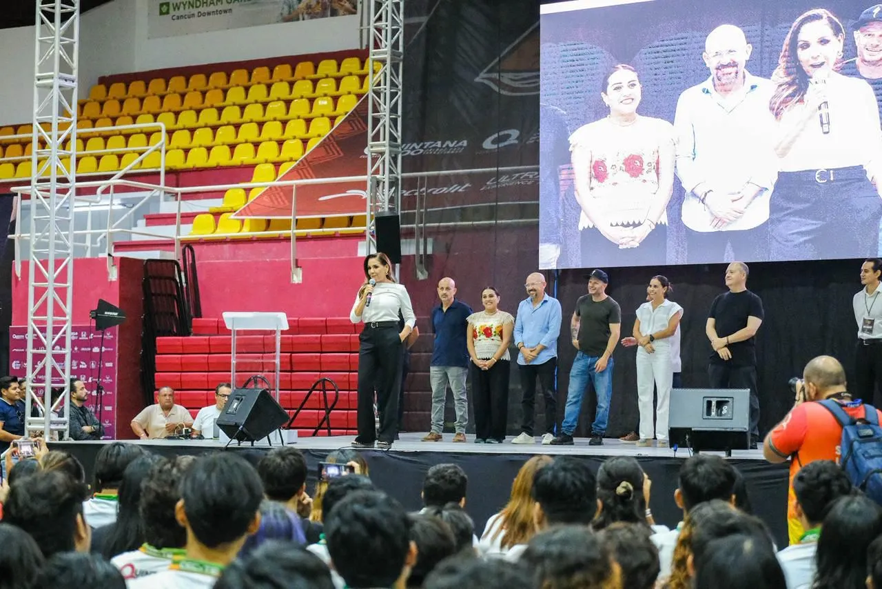 A speaker addressing an audience at an event, with several individuals standing on stage and a large screen displaying a speaker on the side.$#$ CAPTION