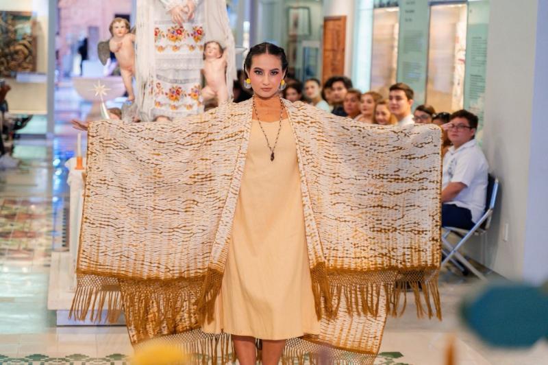 A model wearing a beige dress and a decorative shawl stands confidently, showcasing traditional attire. In the background, an audience watches attentively. A statue and other decorative elements are visible in the setting.