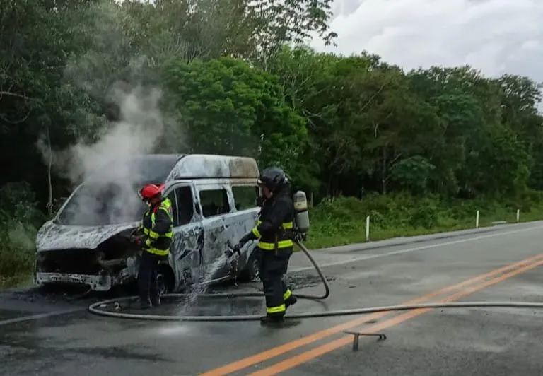 Two firefighters in protective gear are extinguishing a van that is partially burned with smoke rising from it on the side of the road.$#$ CAPTION