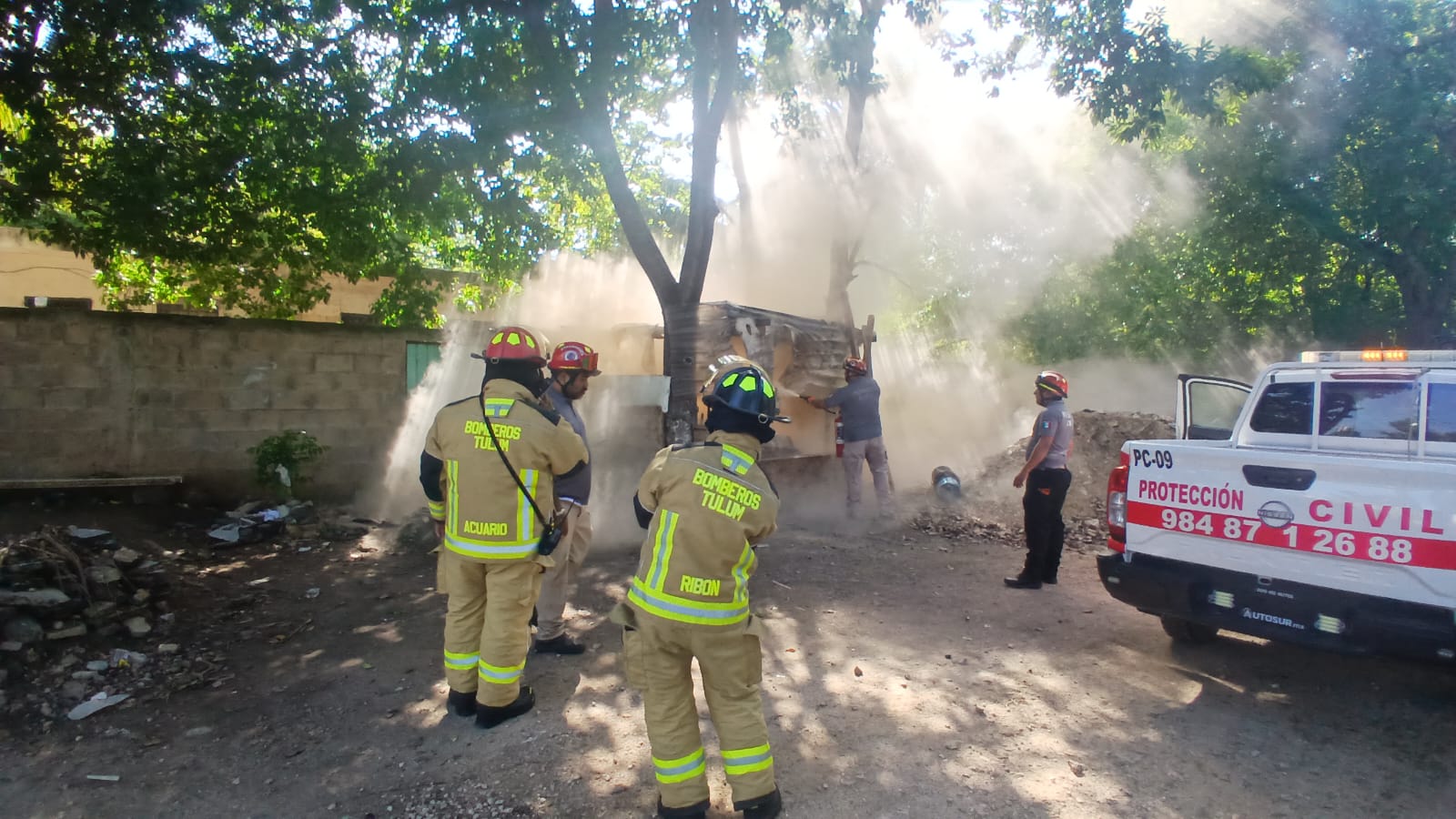 Firefighters working to control a smoke-producing incident in Tulum, Mexico, with a civil protection vehicle nearby. The scene shows emergency responders in action amidst a cloud of smoke.