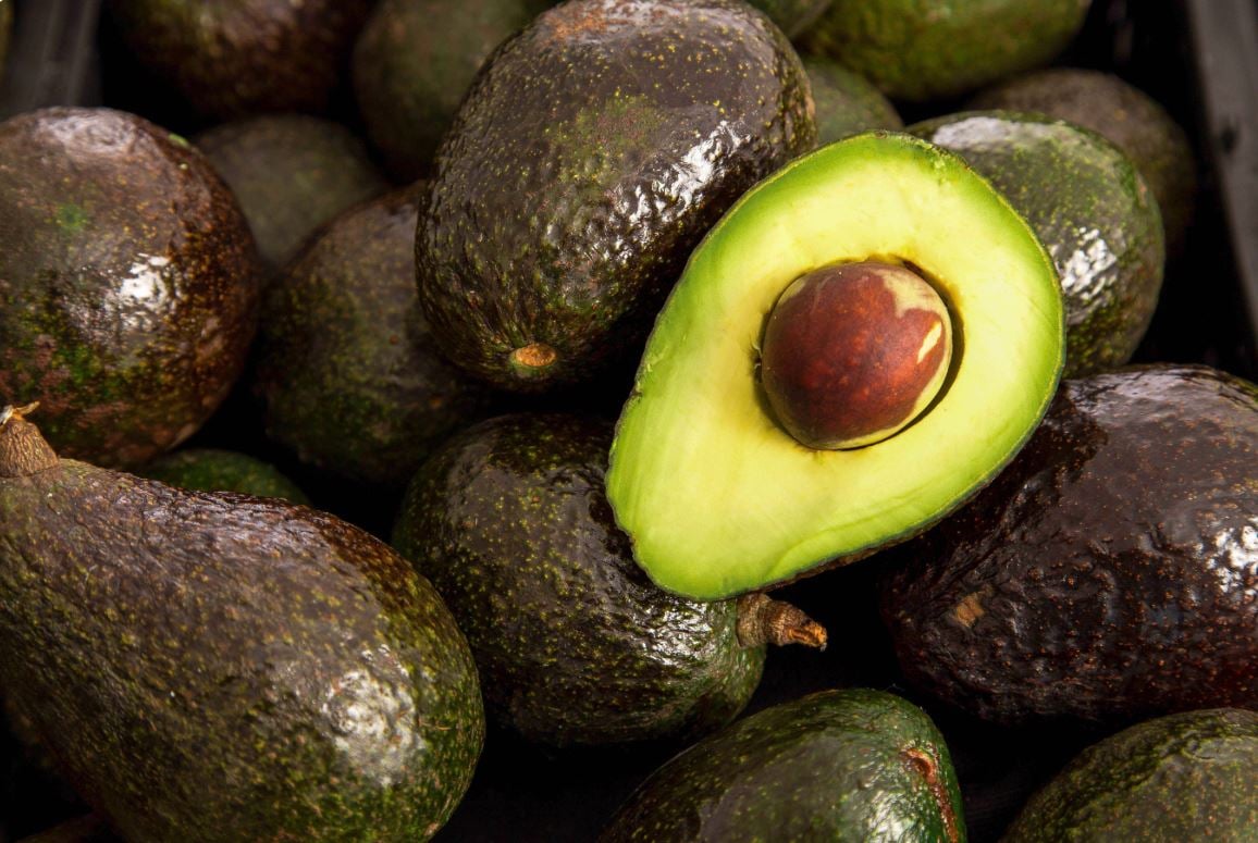 a halved avocado with a dark brown pit surrounded by whole avocados in the background