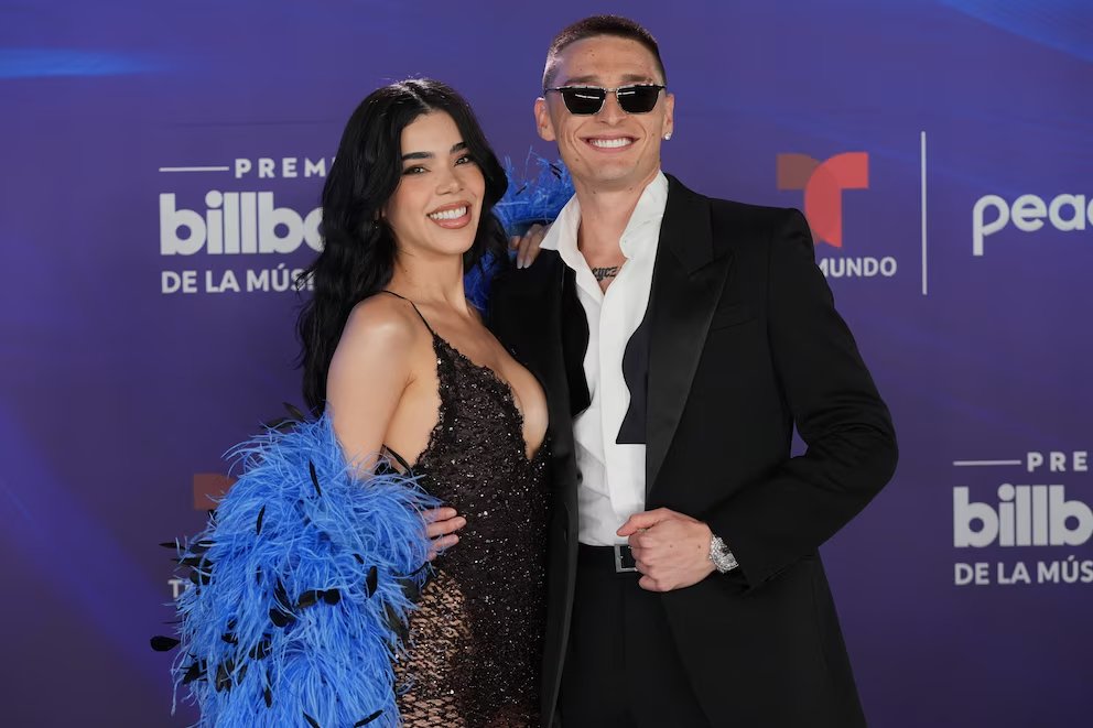A stylish couple poses together at the Billboard Awards, with one wearing a glamorous dress and feathered shawl, and the other in a sleek suit and sunglasses.