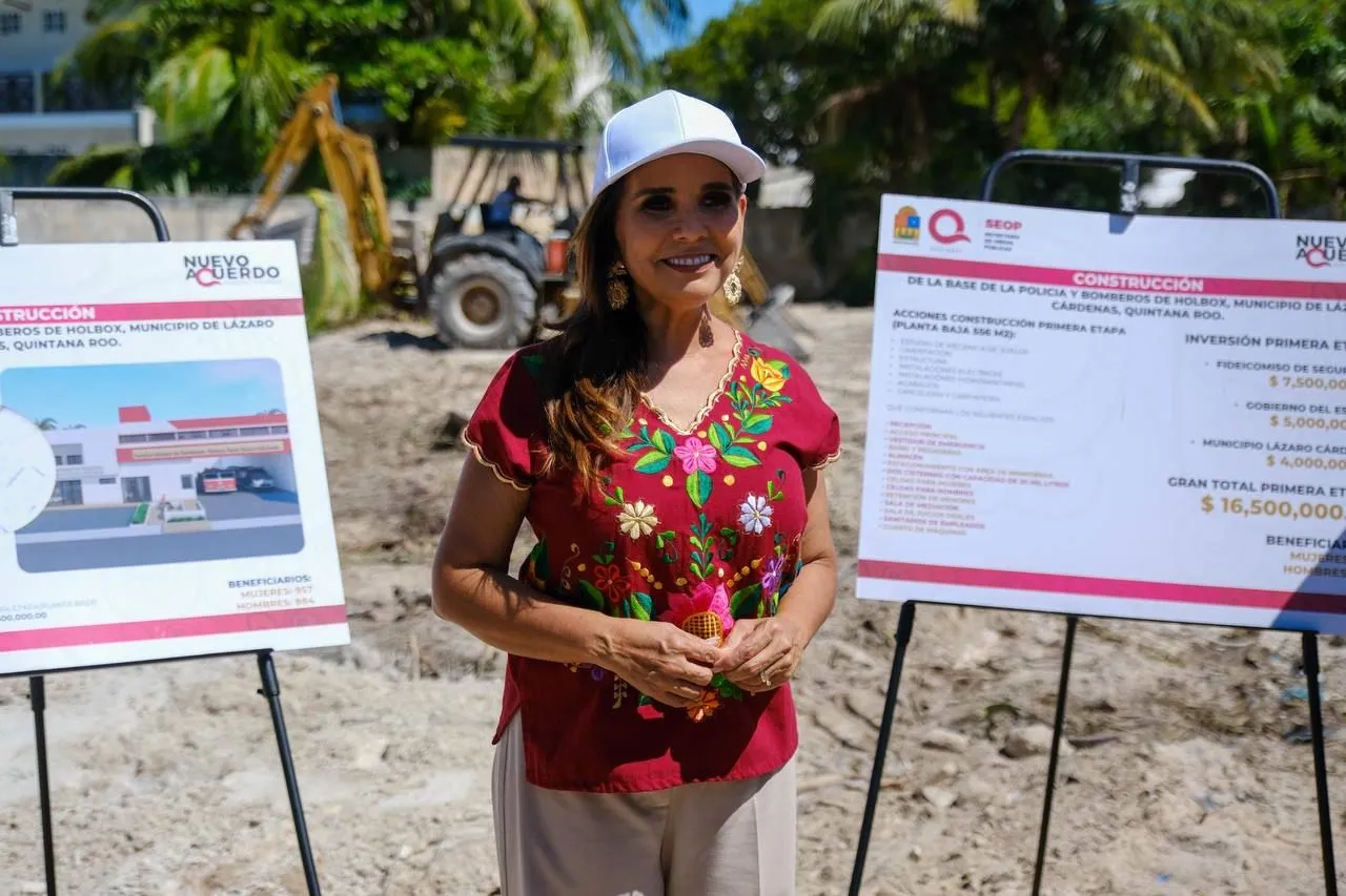 A woman in a colorful traditional shirt stands in front of construction signs at a groundbreaking event in Holbox, Quintana Roo. A tractor is visible in the background.