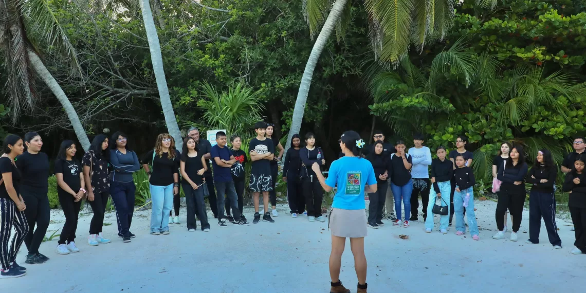 A group of young people in casual clothing gathers on a beach, listening attentively to a speaker wearing a blue shirt. Tall palm trees and lush greenery surround them.$# CAPTION